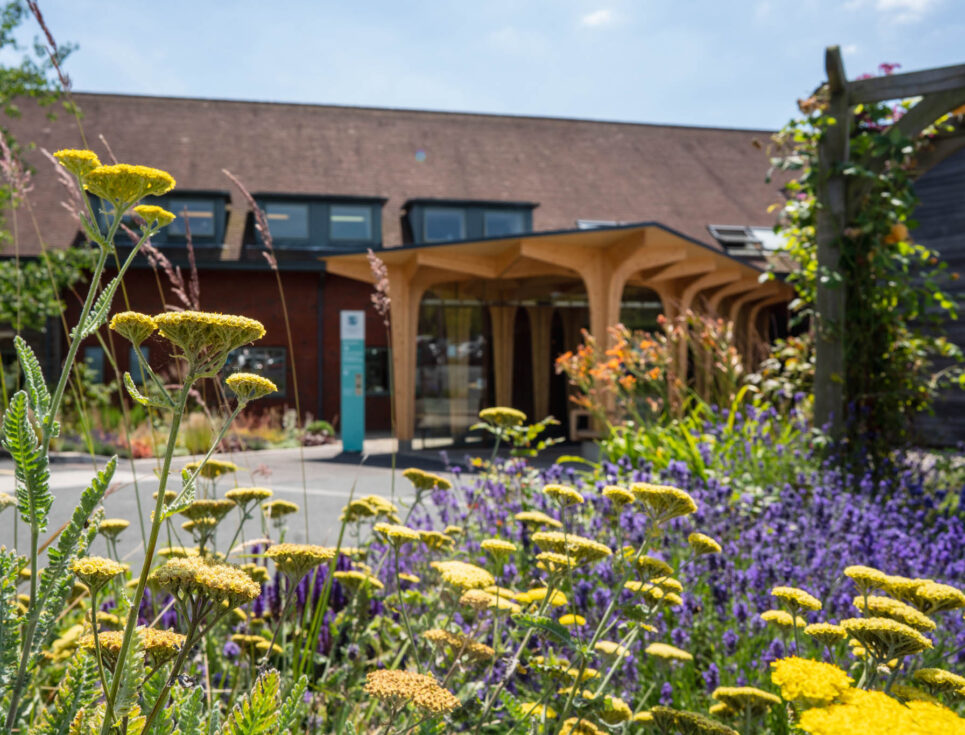 A photograph showing the front entrance to St Richard's Hospice in Worcester. There are yellow and purple flowers in the foreground.