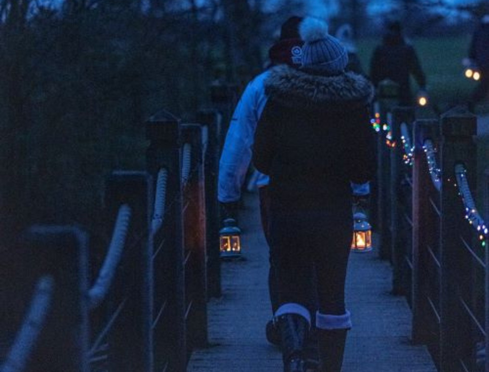 A woman walking over a footbridge at dusk with a lantern in hand.