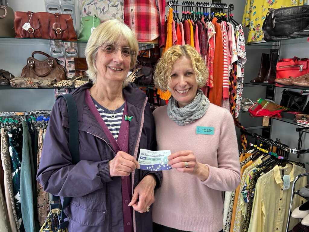 Two women standing in a St Richard's charity shop, both holding the end of a raffle ticket.