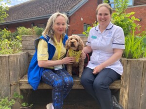 Trisha, sits on a garden bench with Pets as Therapy dog Luna and Therapy Team Leader Alex