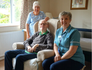 Carole stands behind an armchair in which her husband, Larry is sitting. Senior Healthcare Assistant, June, sits on a chair beside Larry. June is wearing a blue uniform. Everyone is smiling.