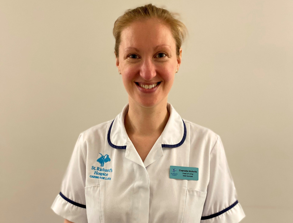 Charlotte stands in front of a plain white wall wearing her physiotherapist uniform. She is smiling.