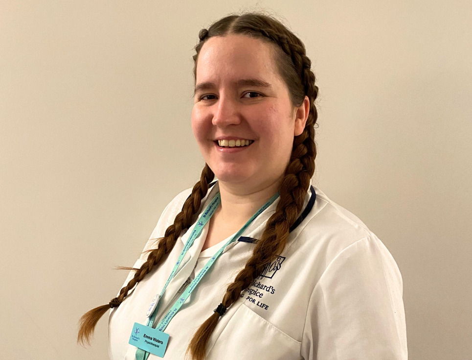 Emma stands in front of a white wall, smiling. She is wearing her white physiotherapist's uniform and her hair is in two long French plaits.