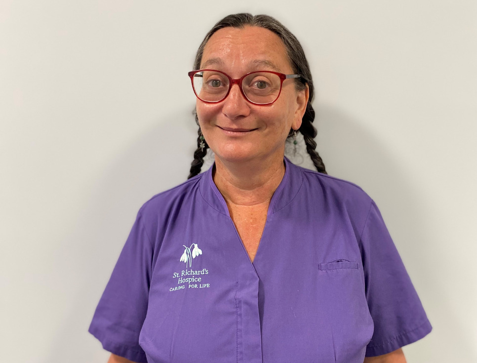 Mary pictured in front of a plain white wall. She is wearing a purple tunic uniform top and wears her hair in two long plaits. She has red-framed glasses on and is smiling.