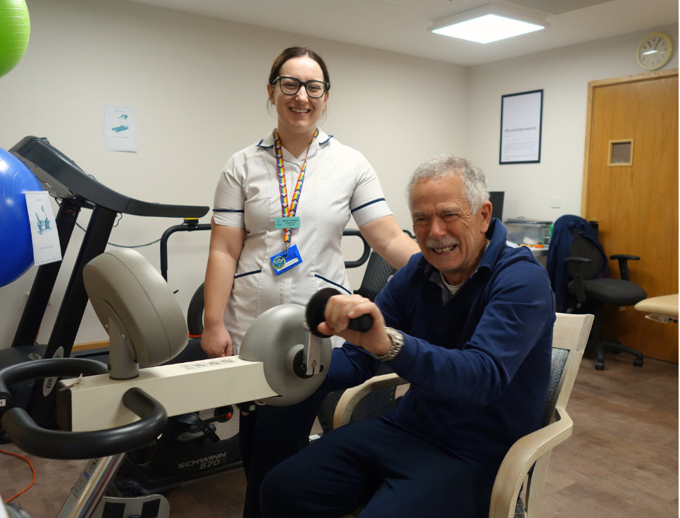 A physiotherapist stands with a patient who is sat using an exercise bike. They are in the hospice's physiotherapy gym.