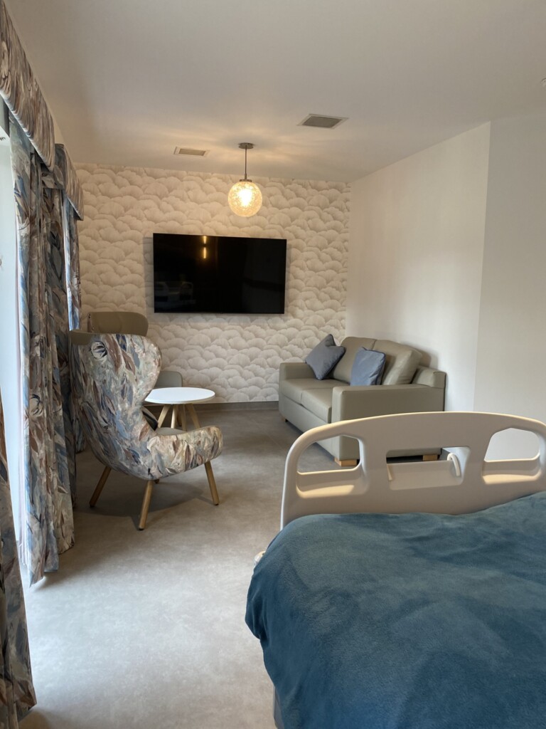 An area of a patient's bedroom in the hospice's In-patient Unit. Comfy chairs are placed around a wall-mounted flat screen TV.