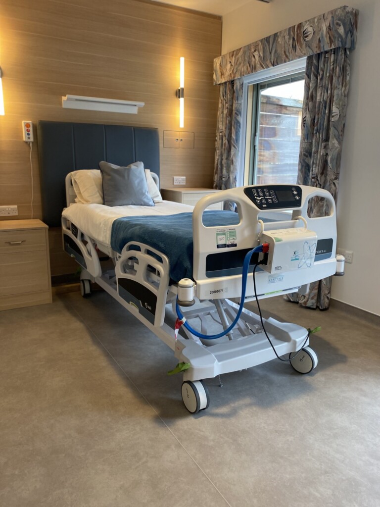 A patient bed in a bedroom on the hospice's In-patient Unit. Next to the bed is a large window looking out onto the gardens. 