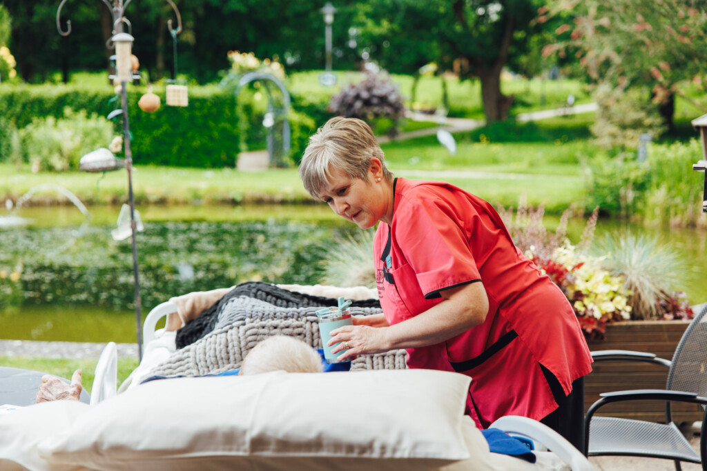 A member of hospice staff in a pink uniform hands a patient a drink in bed. The patient's bed has been taken out onto the patio so he can look out across the gardens. 