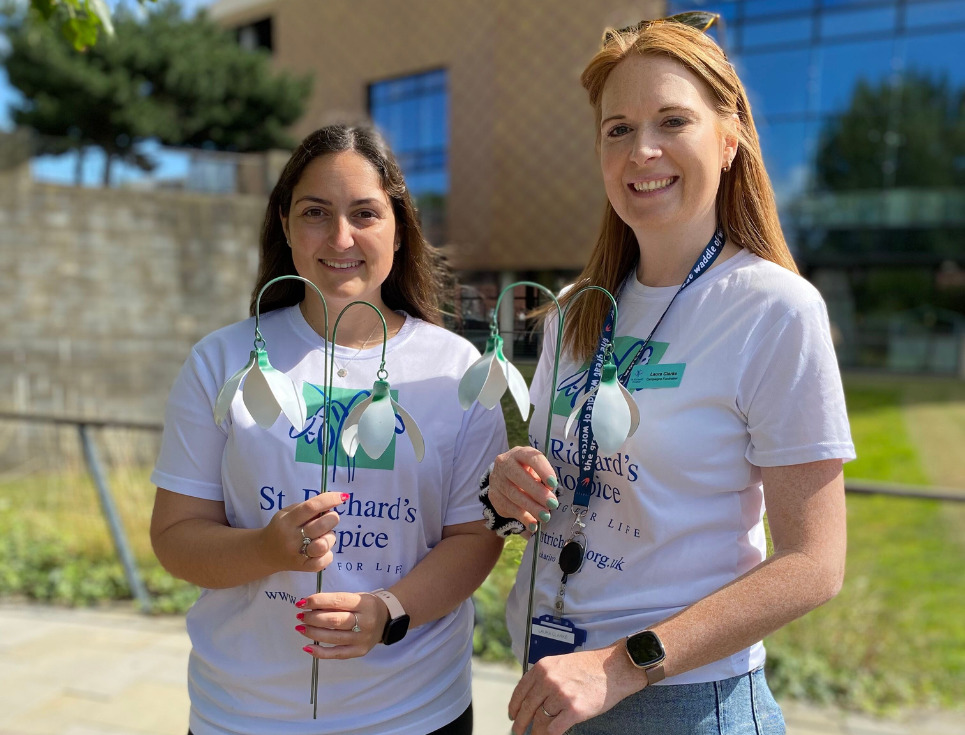 Kelly and Laura stand side-by-side outside the golden clad Hive library holding metal snowdrop sculptures. They are both wearing St Richard's branded t-shirts and smiling.