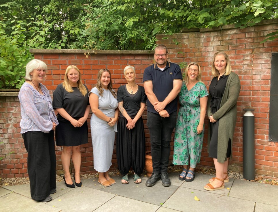 A group of 7 members of the Social Work team stand together with a backdrop of a redbrick wall and greenery.