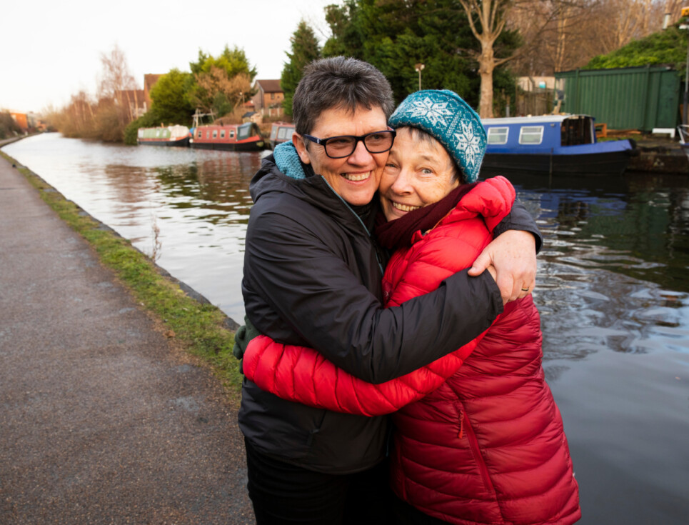 Two people embracing in a hug, smiling. They are both dressed in warm coats and are standing by a canal.