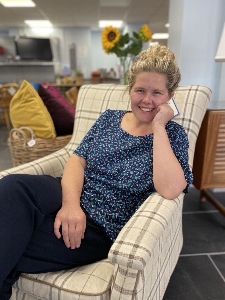 Chloe sits on a comfy armchair in a hospice shop. She is wearing her nurses uniform.