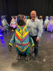 A man wearing jeans and a blue shirt stands smiling next to a penguin sculpture decorated to look like it's wearing an eighties tracksuit. 