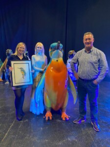 Three people stand with a large penguin sculpture painted to look like a kingfisher. One person is holding a framed painting of a kingfisher. Everyone is smiling. They are at the Waddle of Worcester Auction event.