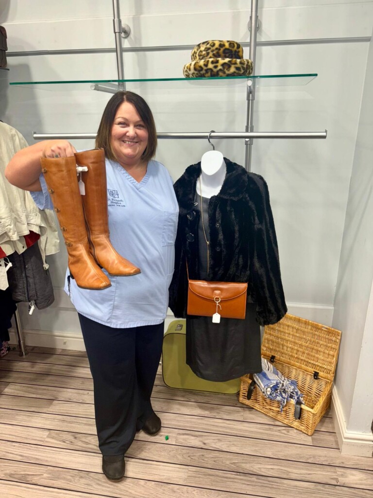 Hayley stood holding an outfit in a hospice shop. She is smiling and wearing her healthcare assistant uniform.