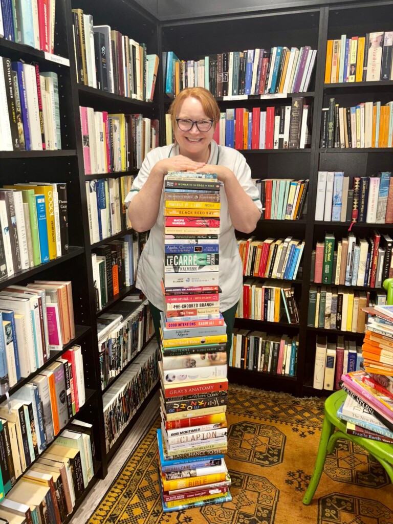 Sally pictured with a stack of books in the hospice's book department. She is stood behind the stack, resting her chin on the top book and smiling.