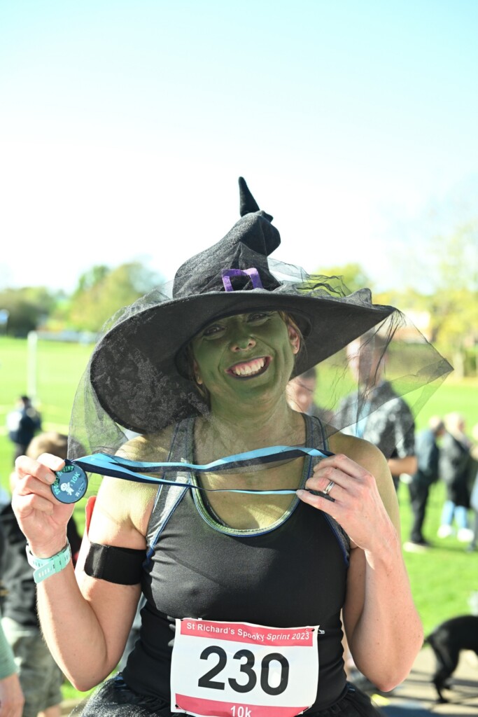 A woman dressed in running gear, a witches hat, and with a green painted face stands smiling with her Spooky Sprint medal.