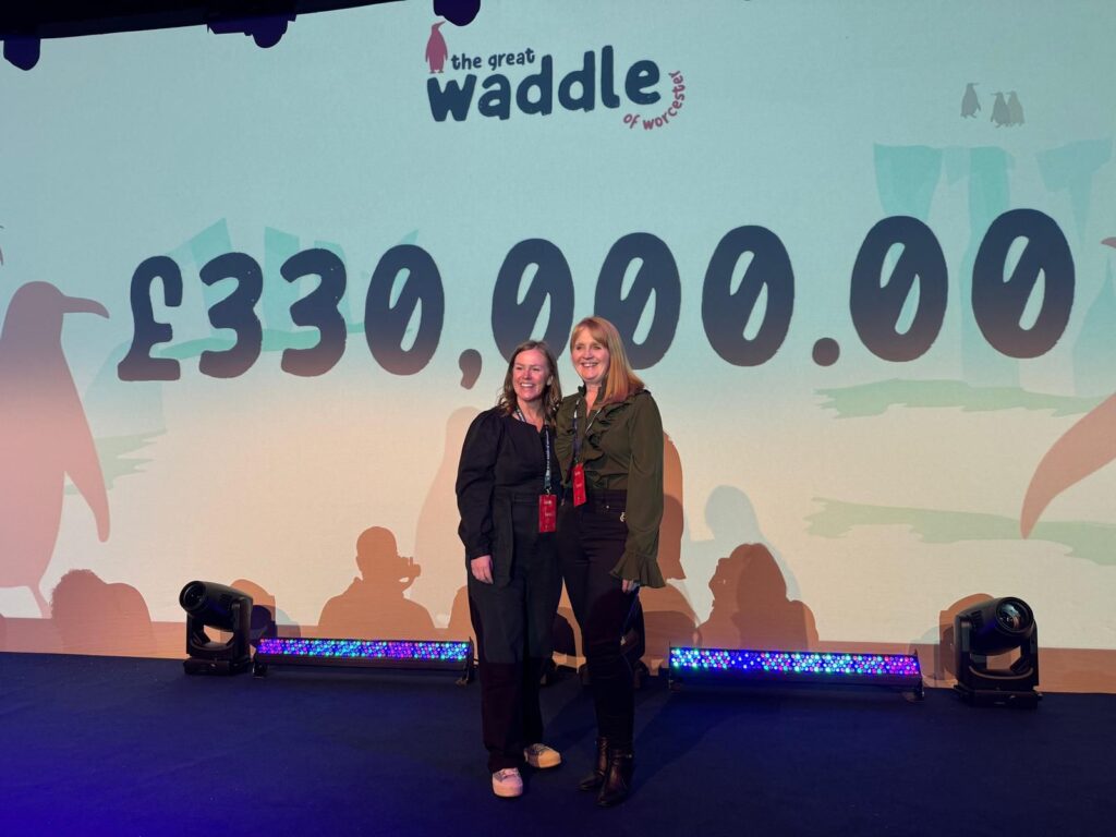 Two women stand on stage in front of a large screen with the Waddle of Worcester auction total of £330,000 emblazoned across it.