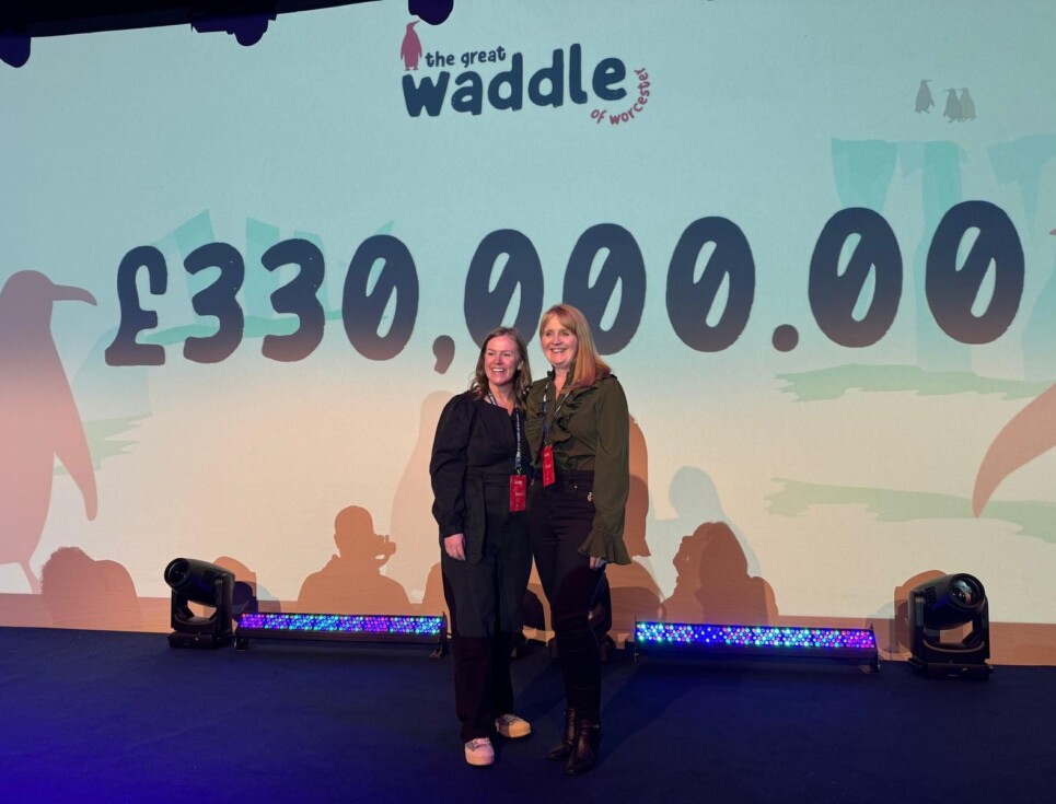 Two women stand on stage in front of a large screen with the Waddle of Worcester auction total of £330,000 emblazoned across it.