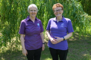 Chaplains Clare and Sarah stand side-by-side in the hospice garden. Behind them is a vibrant green tree. They are both wearing purple tops featuring the hospice logo, and black trousers. They are both smiling.