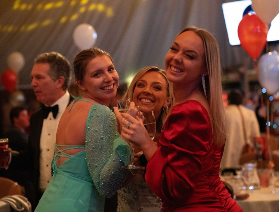 Three women stand together smiling at the Snowdrop Ball. They are all wearing eveningwear dresses.