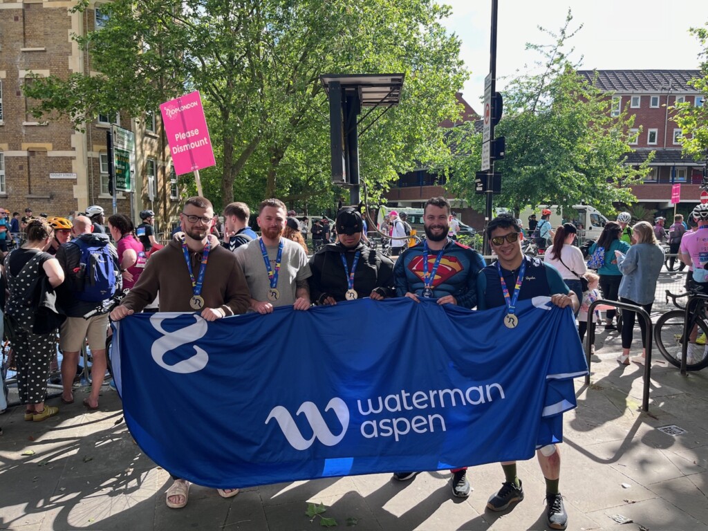 Four people stand together after completing the Ride London challenge. They are holding a blue banner with the Waterman Aspen branding emblazoned on it. Two of the group are in superhero fancy dress.