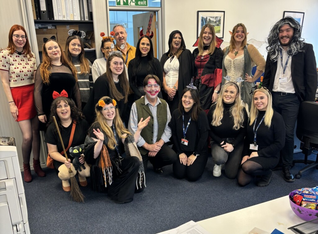 A group of people dressed in Halloween fancy dress in an office. Everyone is smiling.