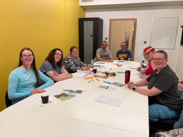 A group of people sat around a big table having a meeting. Everyone is looking at the camera and smiling. The walls are painted yellow and white. 