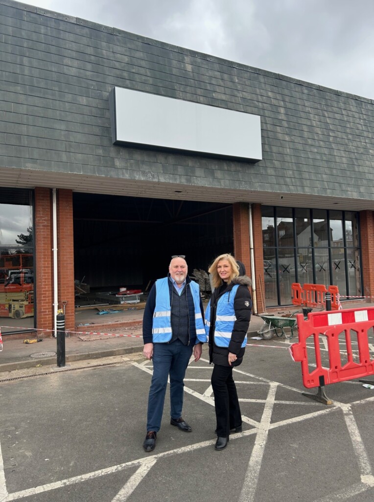 Dan and Abby from the hospice's income generation team stand outside the superstore which is undergoing building work. They are both wearing warm clothing and blue high-vis jackets.