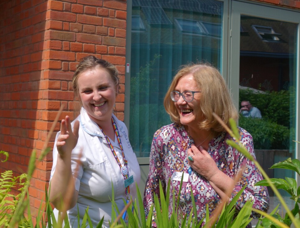 Samantha, pictured in white therapy support assistant uniform, stands laughing and smiling with Sue in the courtyard garden at St Richard's in the sunshine. Sue is smiling and wearing a purple patterned dress.