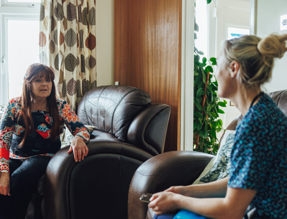 A nurse speaks to a patient in the patient's front room at home.