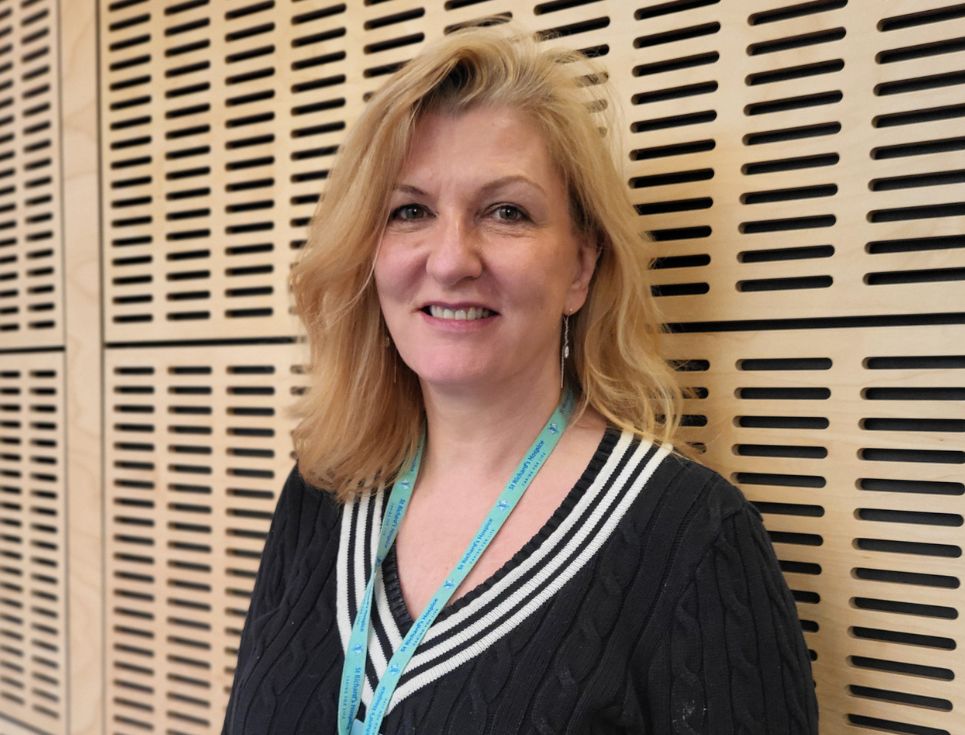 Abby pictured standing in front of a wood-panelled wall at the hospice. She has shoulder-length blonde hair and is wearing a black and white jumper.