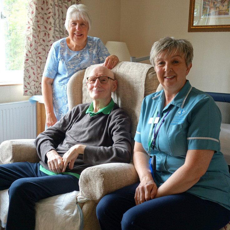 Carole stands behind an armchair in which her husband, Larry is sitting. Senior Healthcare Assistant, June, sits on a chair beside Larry. June is wearing a blue uniform. Everyone is smiling.