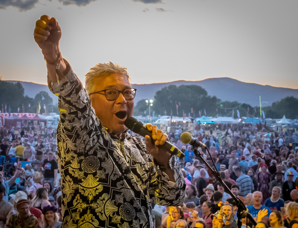 Person on an outdoor stage at a music festival holding a microphone and raising a fist, with a large crowd and festival tents in the background at dusk.