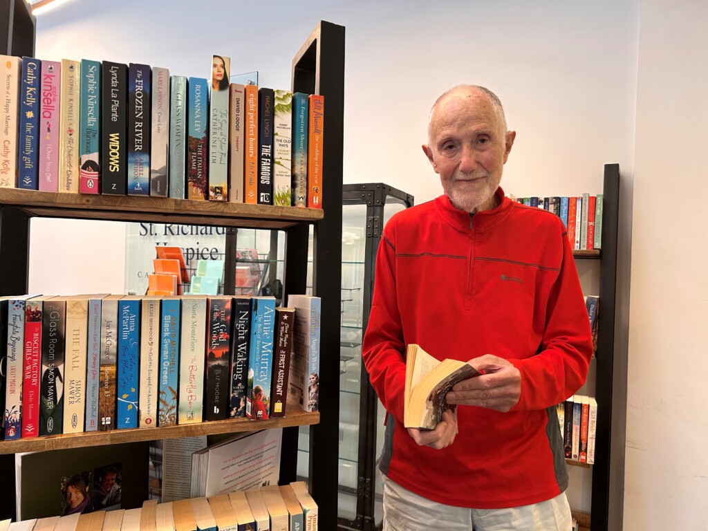 Person in a red jumper stands beside wooden bookshelves filled with novels, holding an open paperback book. Behind the shelves, a sign with the words “St Richard’s Hospice” is partly visible..