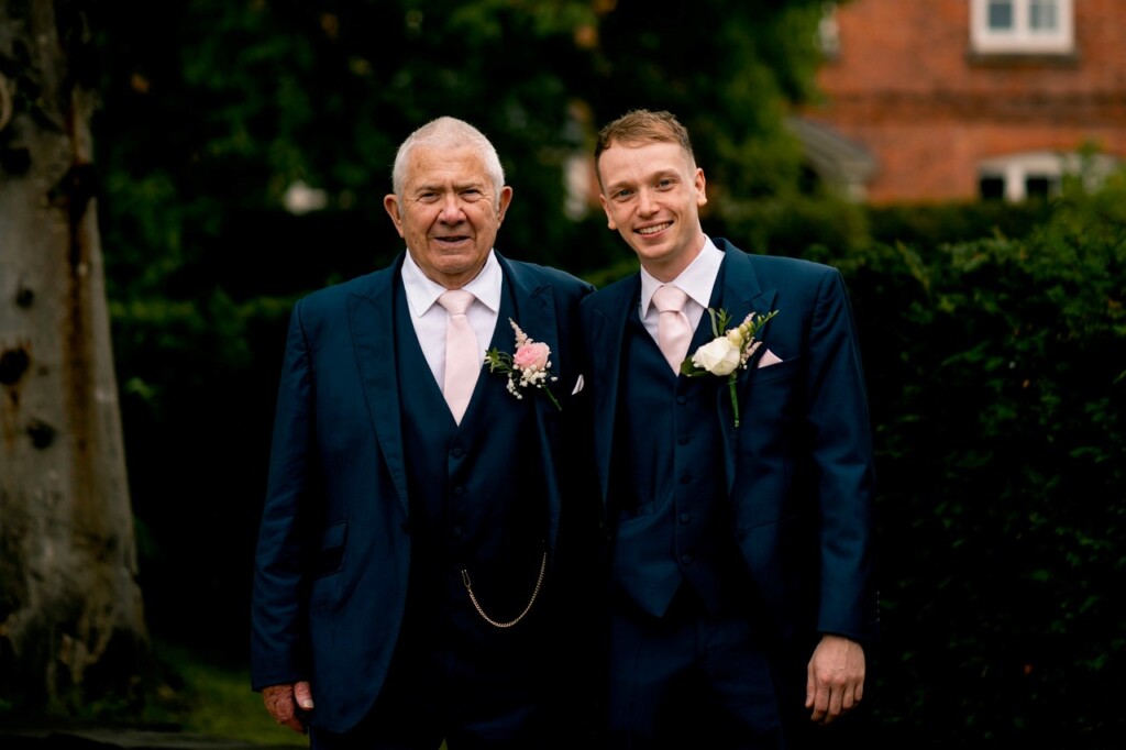 A grandfather and grandson wearing dark suits with pale pink ties and buttonhole flowers stand side by side outdoors, with trees and a brick building in the background.