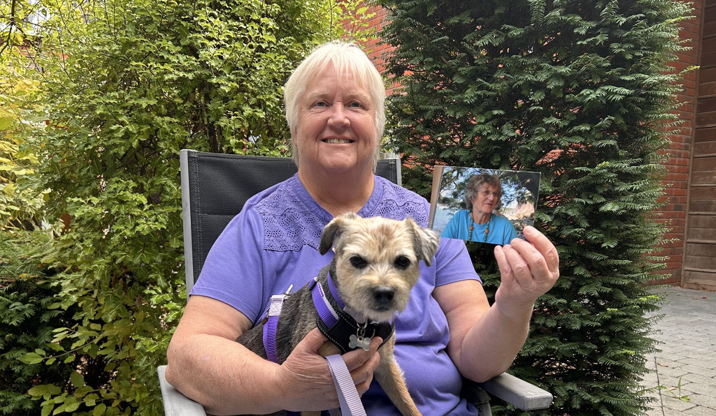 Person sitting outdoors with a small dog on their lap, holding a photograph of another person. The image is taken in a garden setting with trees and greenery in the background.