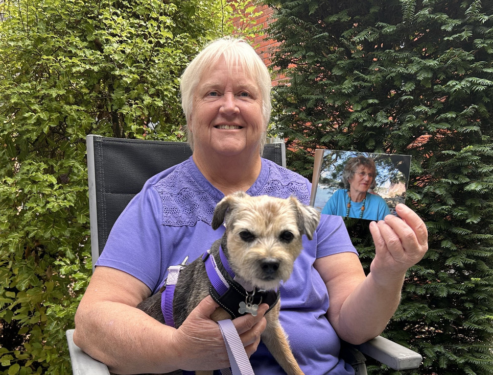 Person sitting outdoors with a small dog on their lap, holding a photograph of another person. The image is taken in a garden setting with trees and greenery in the background.