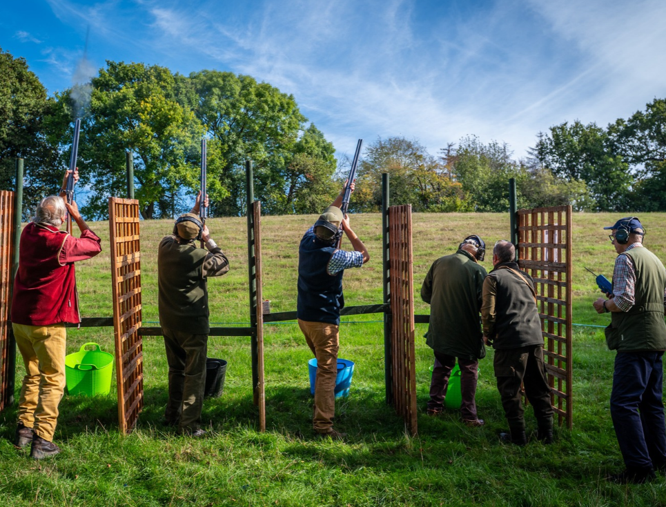 Clay pigeon shooting participants stand at outdoor shooting stations, aiming shotguns towards targets across a grassy field.