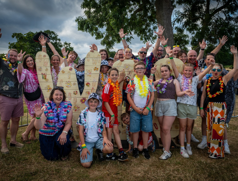 Group of adults and children wearing colourful leis pose in front of Woo Fest sign at an outdoor summer music festival.